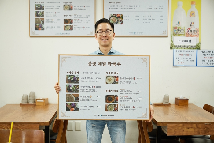 서울시 양천구에 위치한 '봉평 메밀 막국수' 사장님이 바뀐 메뉴판을 들고 포즈를 취하고 있다. 〈자료 우아한형제들〉