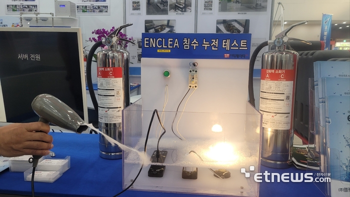 티제이티플러스가  전시회 현장 부스에서 엔클리어의 소화약제 용액을 담근 전기제품을 작동하며 누전시연을 하고 있다.