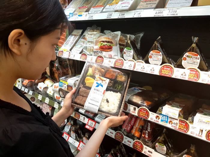 고객이 이마트24 맛보장 홍보물이 붙어 있는 FRESH FOOD 매대에서 맛보장 도시락을 살펴보고 있다.